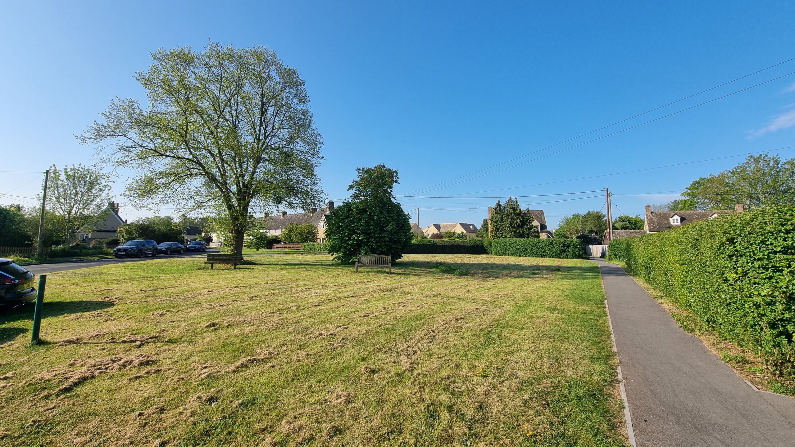 Full image of A village green with trees and benches under clear blue skies and Spring morning sunlight.