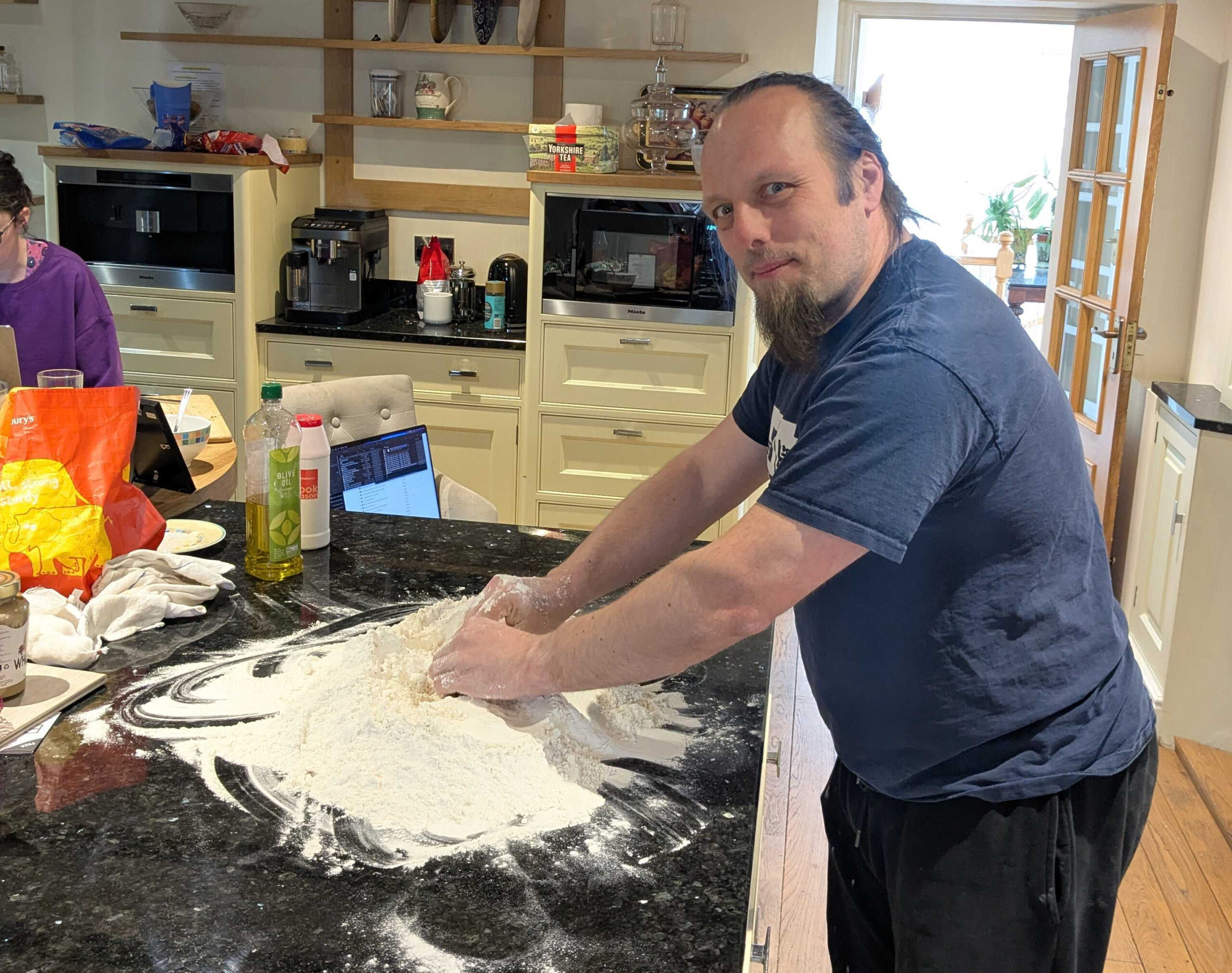 Full image of Dan, a white man with a ponytail of blue hair and a goatee beard, uses his hands to gather a huge pile of flour on a marble worksurface in a spacious kitchen.
