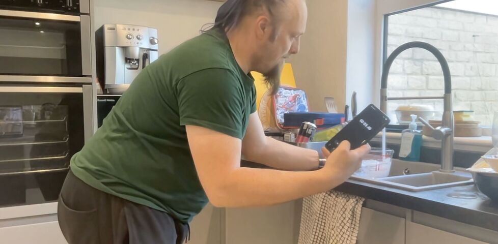 Dan, a white man with a blue ponytail, wearing a green t-shirt, watches a stopwatch timer on his phone while filling a measuring jug from the kitchen sink.