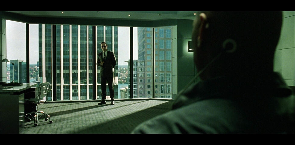 Framegrab from The Matrix. In the foreground is the silhouette of Morpheus, who is about to be interrogated by Agent Smith, a man in a suit at the windowed far end of an office.
