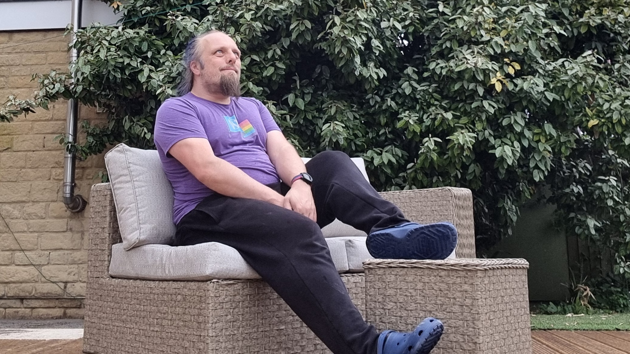 Full image of Dan, a white man with a blue ponytail and a goatee beard, wears purple t-shirt, tracksuit bottoms, and crocs to sit outdoors on garden furniture in light rain.