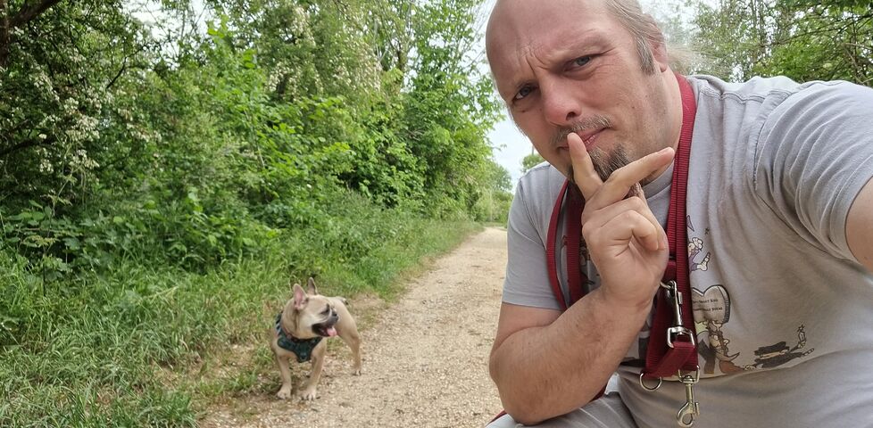 Dan, a white man with a goatee beard and a blue ponytail, looks thoughtful as he crouches on a footpath near a fawn-coloured French Bulldog. 