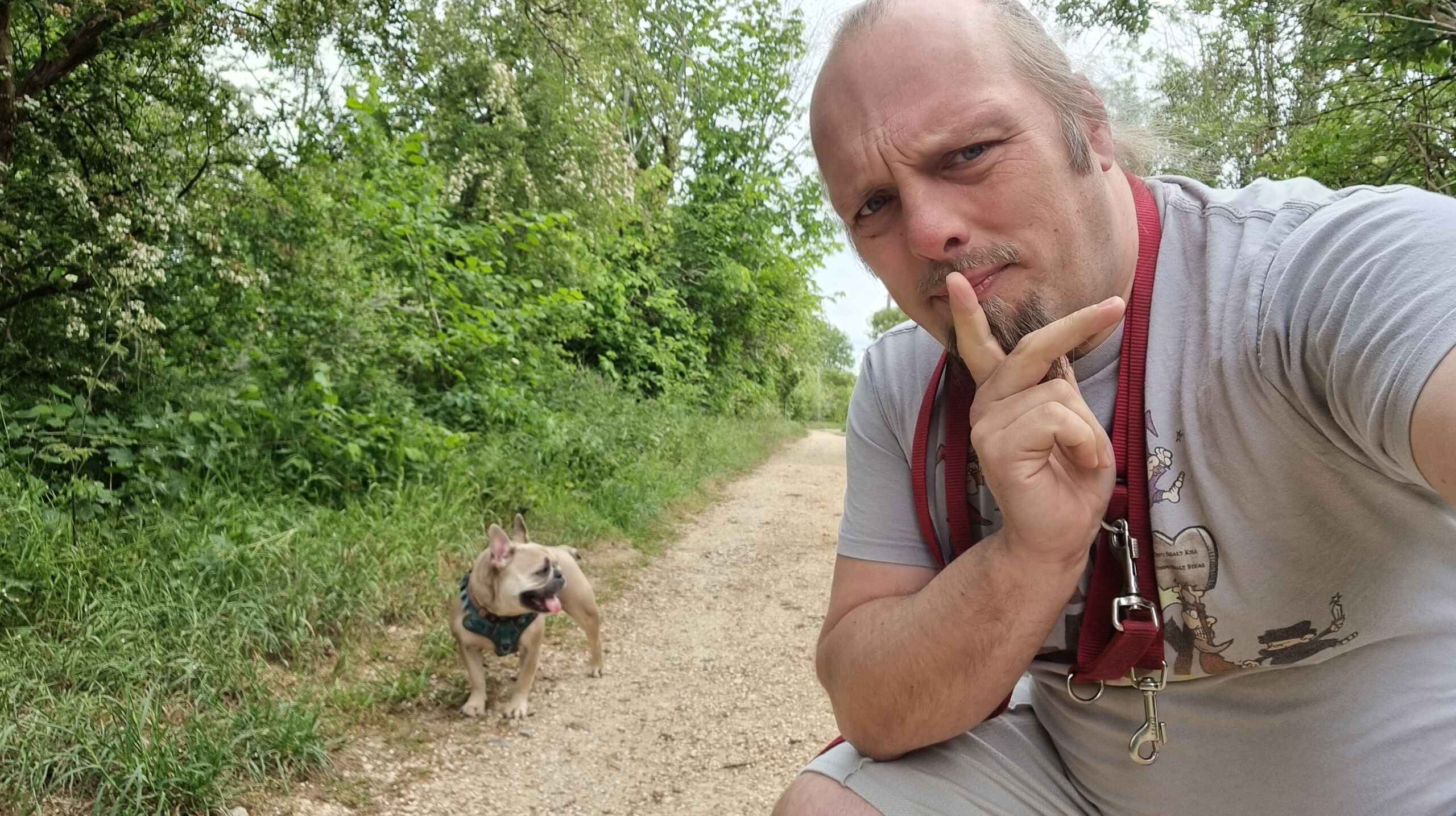 Full image of Dan, a white man with a goatee beard and a blue ponytail, looks thoughtful as he crouches on a footpath near a fawn-coloured French Bulldog.