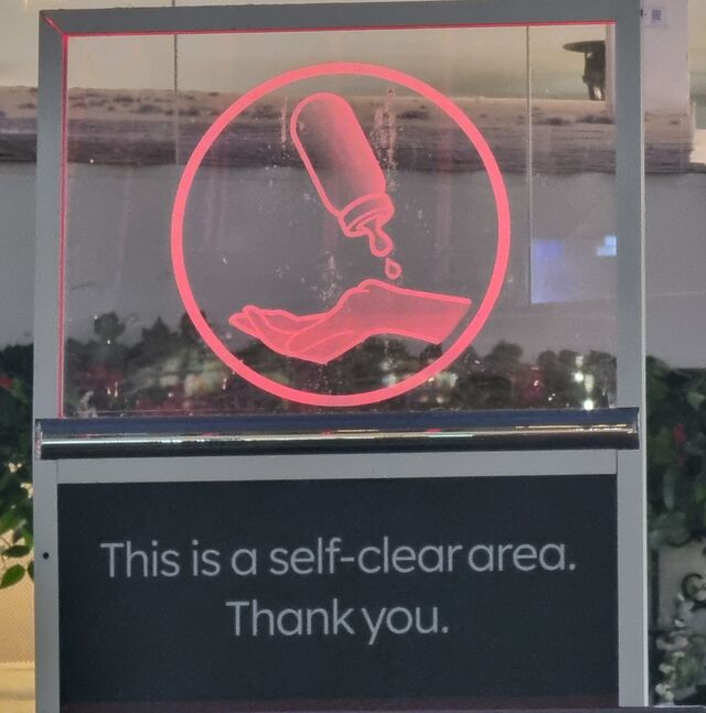 Printed sign reading 'this is a self-clear area; thank you' beneath a red illuminated icon of a hand onto whose wrist a bottle drips.