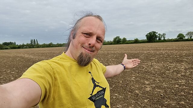 A white man wearing a yellow 'Slamilton' t-shirt stands, smiling, in the middle of an empty field.