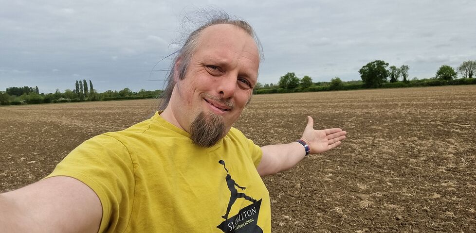 A white man wearing a yellow 'Slamilton' t-shirt stands, smiling, in the middle of an empty field.