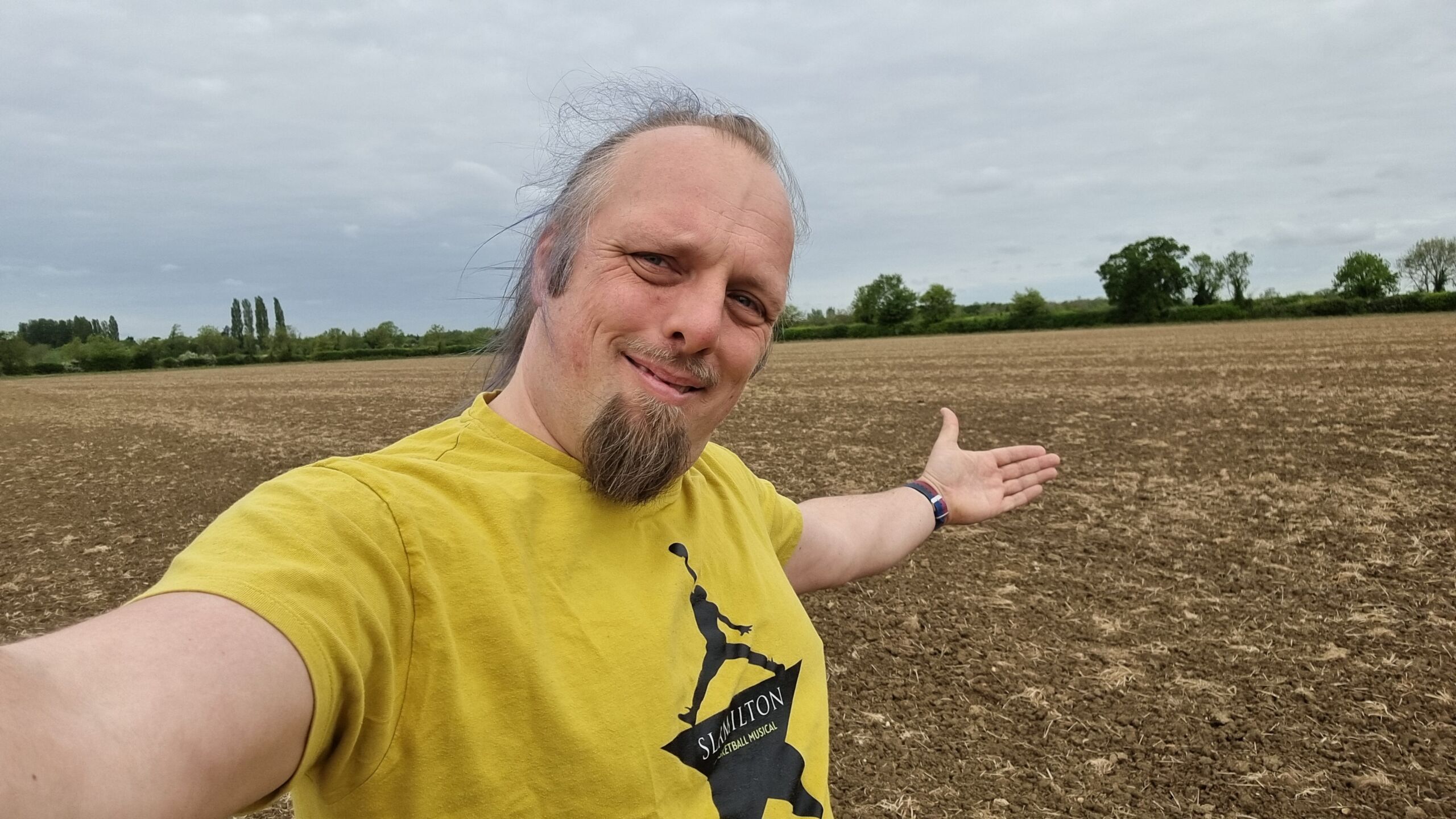 Full image of A white man wearing a yellow 'Slamilton' t-shirt stands, smiling, in the middle of an empty field.