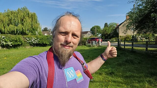 Standing in a field on the outskirts of a rural village, Dan - a white man with a goatee beard , with a dog's lead hung around his shoulders - throws a thumbs-up.