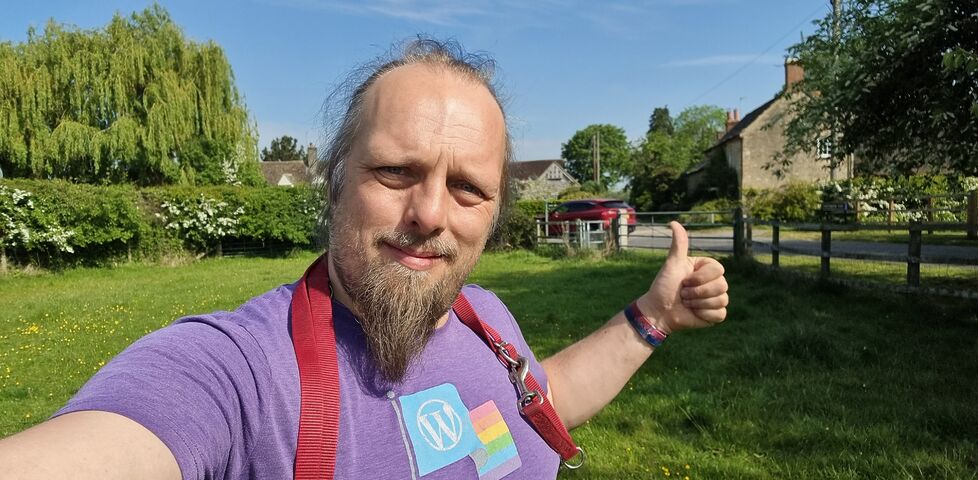 Standing in a field on the outskirts of a rural village, Dan - a white man with a goatee beard , with a dog's lead hung around his shoulders - throws a thumbs-up.