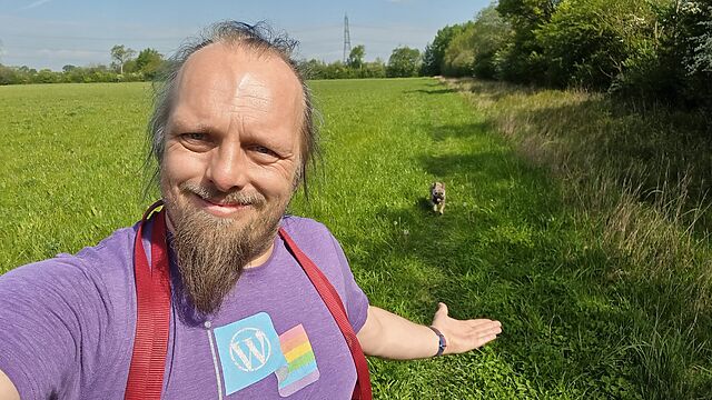Dan, a white man with a goatee beard, smiles as he gestures behind him to the French Bulldog following him through a field.