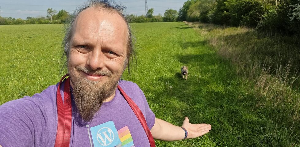 Dan, a white man with a goatee beard, smiles as he gestures behind him to the French Bulldog following him through a field.