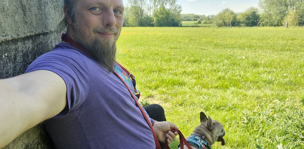 Dan, a white man with a blue ponytail and a goatee, sits in the shadow of an old concrete wall alongside a leashed French Bulldog, with a large meadow visible in the background.