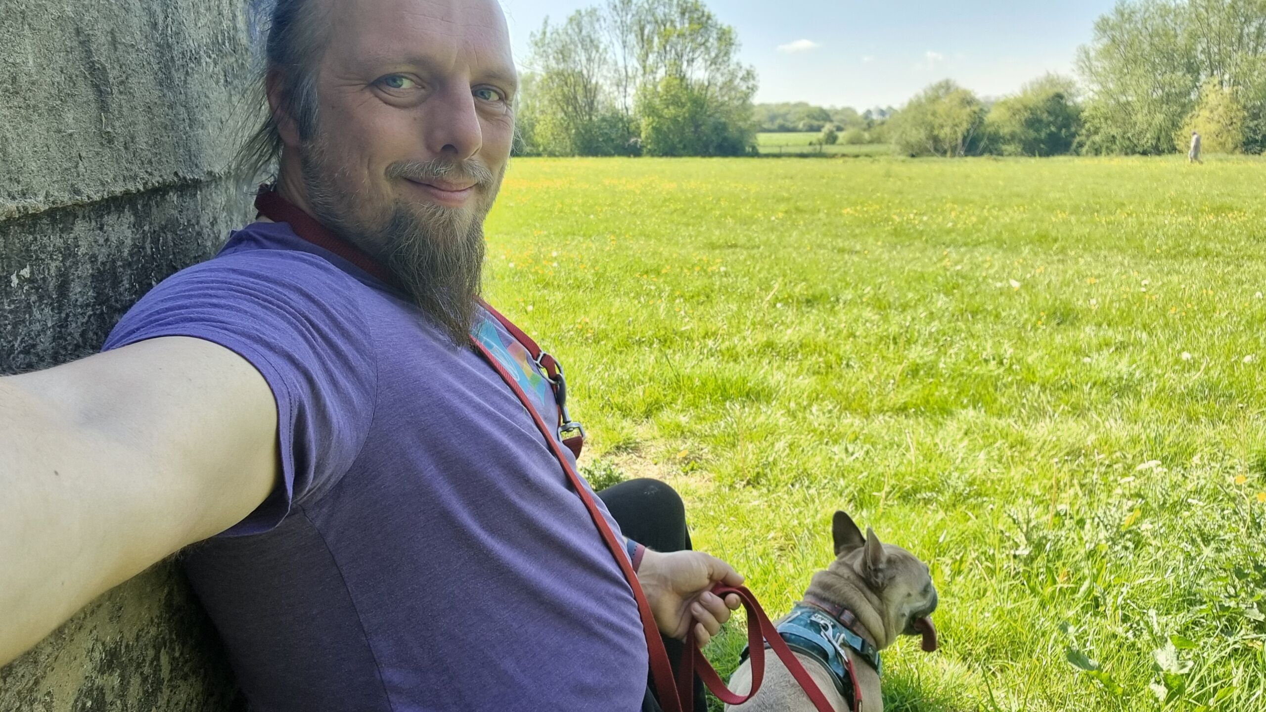 Full image of Dan, a white man with a blue ponytail and a goatee, sits in the shadow of an old concrete wall alongside a leashed French Bulldog, with a large sun-dappled meadow visible in the background.