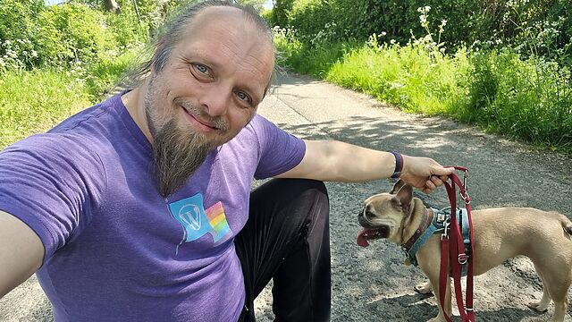 Dan, a white man with a blue ponytail and a goatee beard, wearing a purple t-shirt, crouches in a quiet rural lane alongside a panting French Bulldog, whose lead he's holding, as he smiles at the camera.