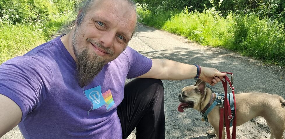 Dan, a white man with a blue ponytail and a goatee beard, wearing a purple t-shirt, crouches in a quiet rural lane alongside a panting French Bulldog, whose lead he's holding, as he smiles at the camera.