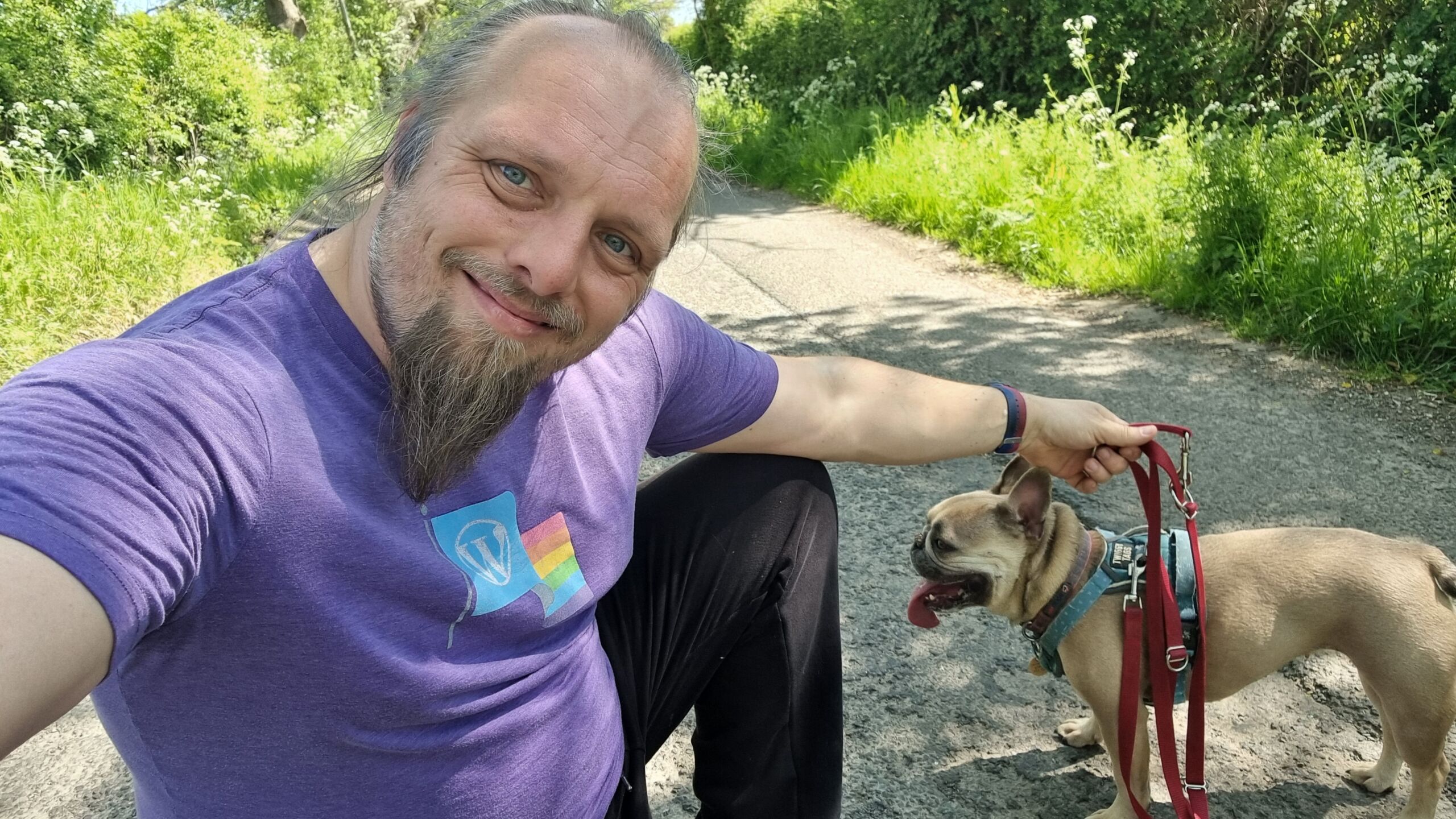 Full image of Dan, a white man with a blue ponytail and a goatee beard, wearing a purple t-shirt, crouches in a quiet rural lane alongside a panting French Bulldog, whose lead he's holding, as he smiles at the camera.
