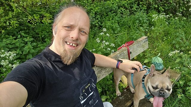Dan, a white man with a goatee beard, sits on a wooden bench in a shady area, alongside a French Bulldog.
