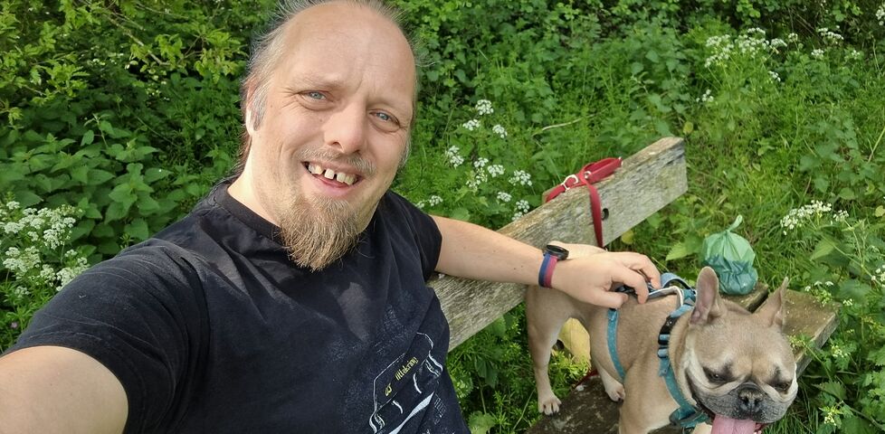 Dan, a white man with a goatee beard, sits on a wooden bench in a shady area, alongside a French Bulldog.