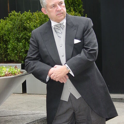 Carl Person, an older white man with receding grey-white hair, wearing a smart three-piece suit, leans against a planter in an marbled ornamental garden.