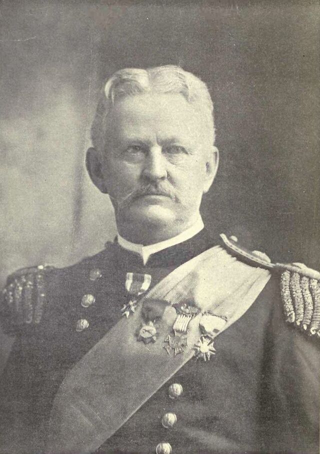 An older whtie man in military uniform with medals and a sash.