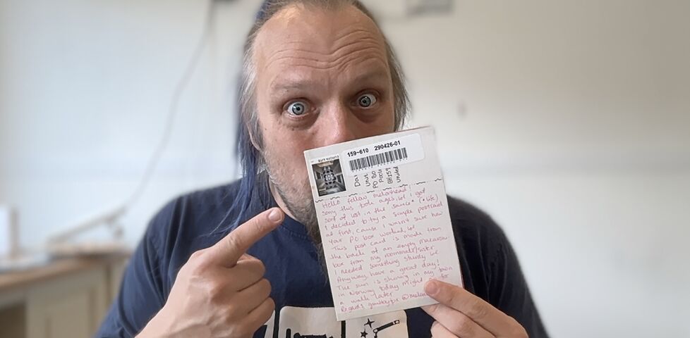 Dan, a white pan with a blue ponytail, holds a postcard in front of his face.