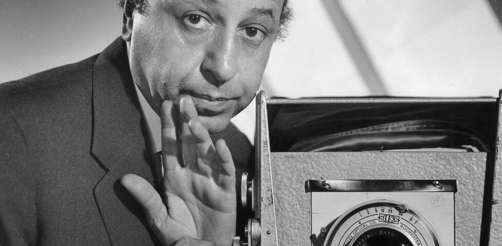 Monochome photo, presumably taken using a mirror, of a balding man operating a box camera.