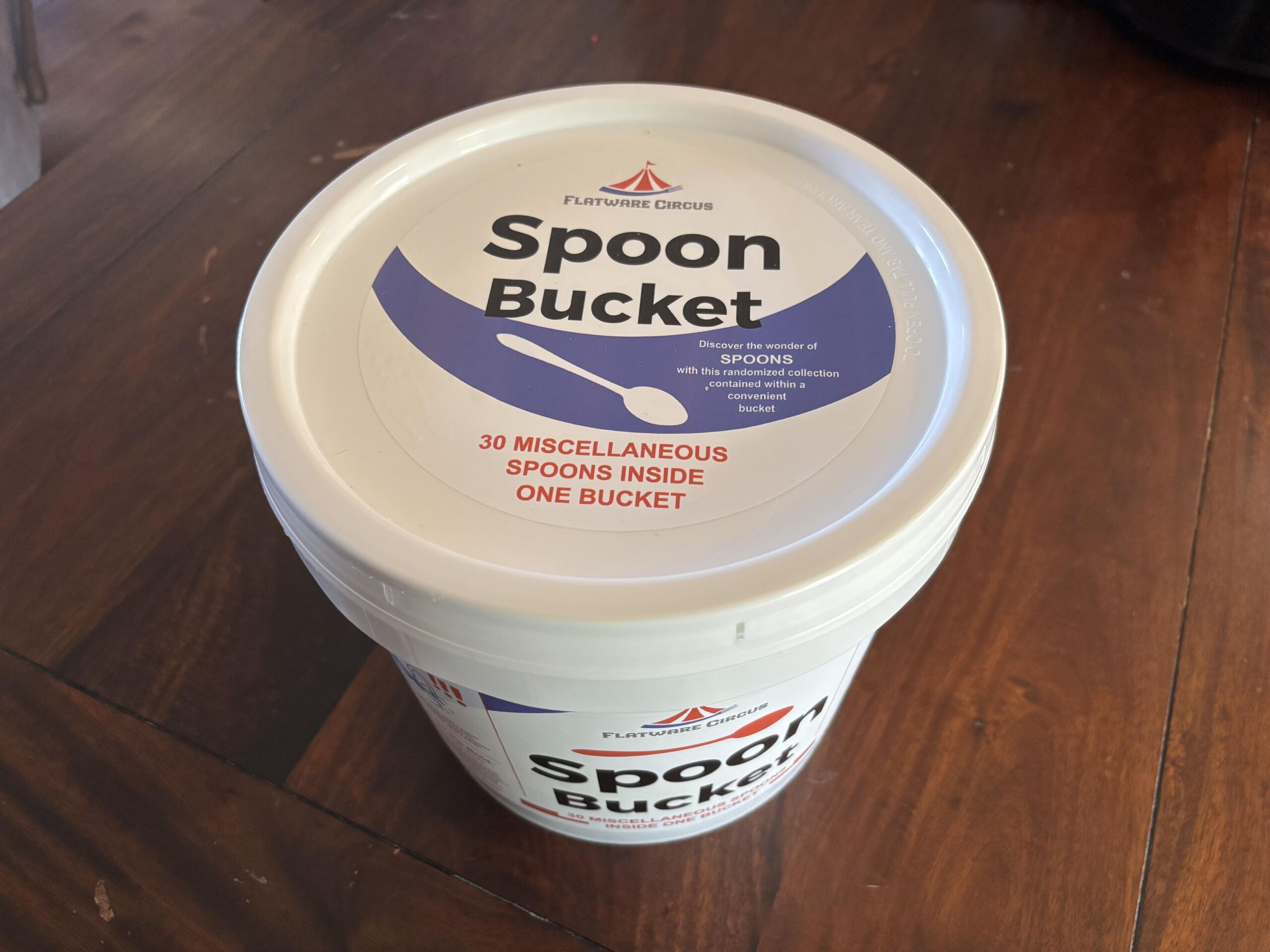 Full image of 2 gallon white plastic bucket, labeled: - Flatware Circus - Spoon Bucket - Discover the wonder of spoons with this randomized collection contained within a convenient bucket - 30 miscellaneous spoons inside one bucket