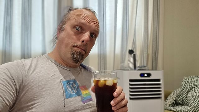 Dan, a white man with a small beard and a blue ponytail, holds a pint if iced coffee on front of a residential portable air conditioner unit.