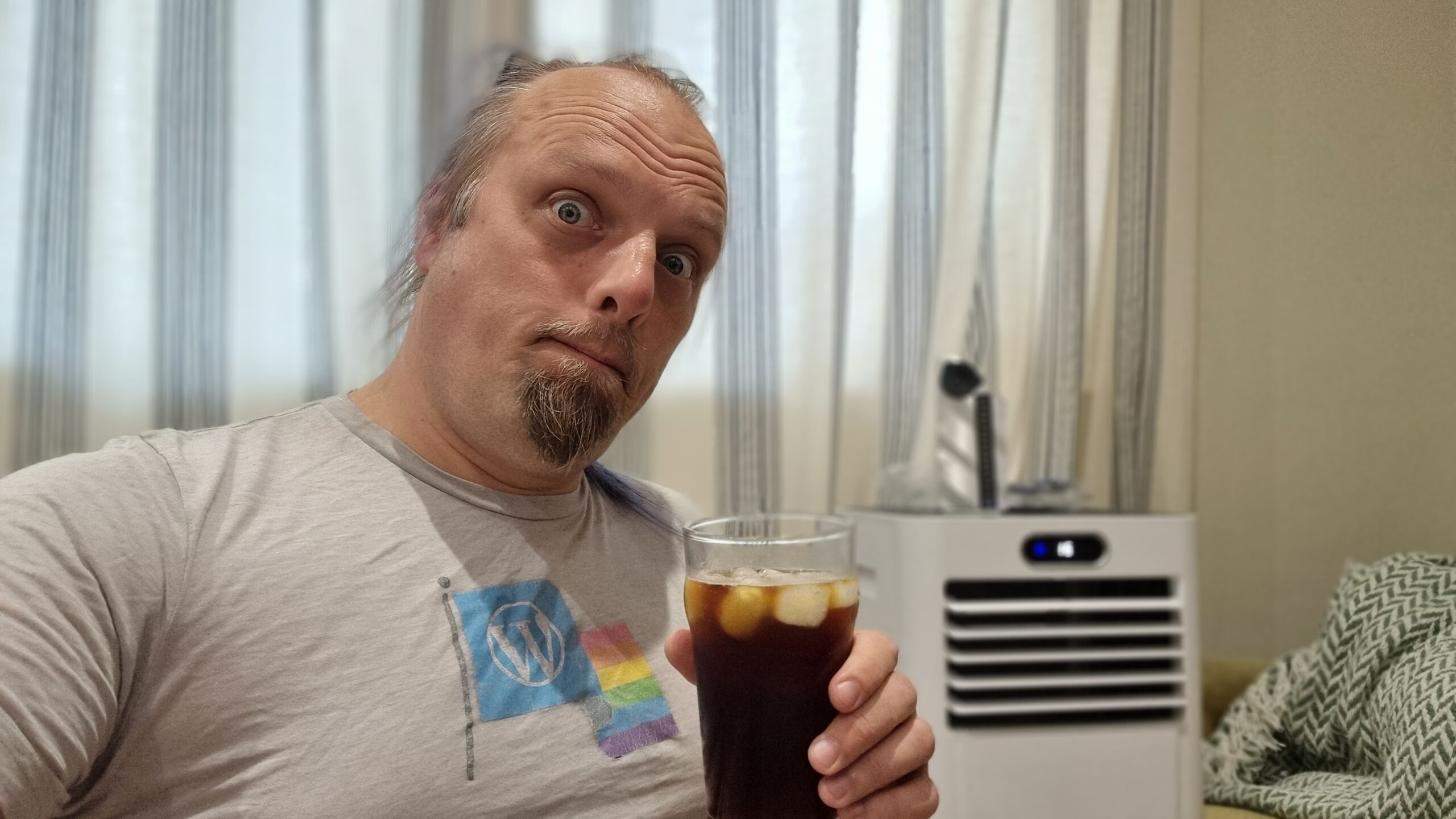 Full image of Dan, a white man with a small beard and a blue ponytail, holds a pint if iced coffee on front of a residential portable air conditioner unit.