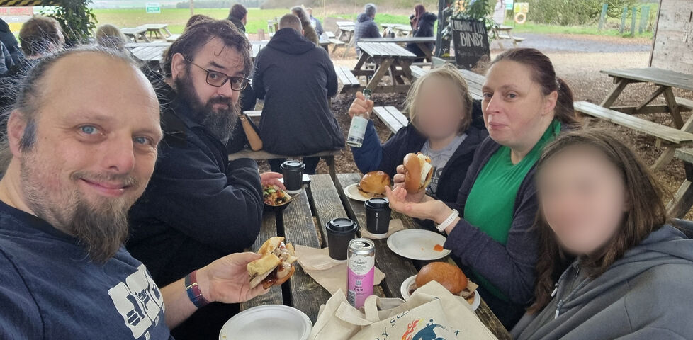A family of two white men with beards and one white woman sit at a picnic bench in a barn, alongside their two (blurred) children. They're holding baps with sausages, bacon, or eggs in, and surrounded by canned drinks and takeaway coffee cups. A tote bag nearby gives away that they're at the cafe of Diddly Squat Farm.
