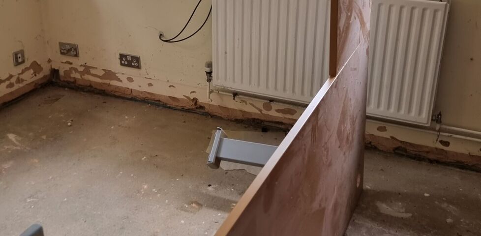 A large, L-shaped office desk with thick motorised metal legs lies on its edge, upright, in a bare concrete-floored home office.