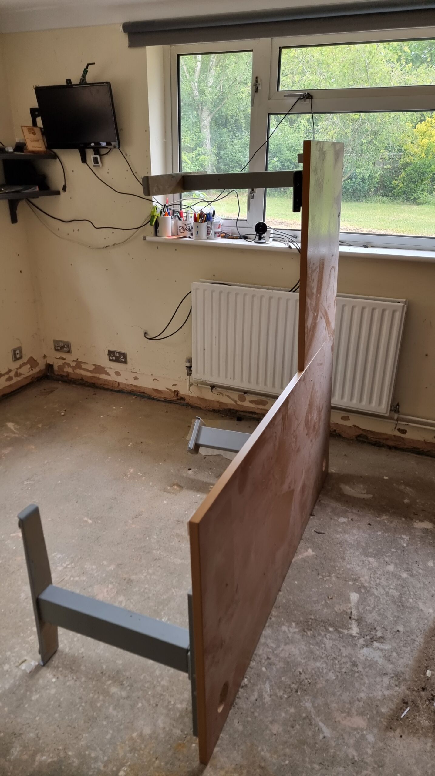 Full image of A large, L-shaped office desk with thick motorised metal legs lies on its edge, upright, in a bare concrete-floored home office.