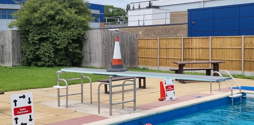 A traffic cone sits atop a diving board at an outdoor swimming pool.