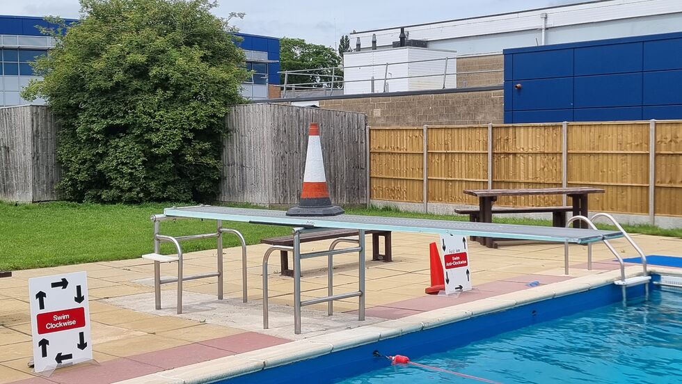 A traffic cone sits atop a diving board at an outdoor swimming pool.