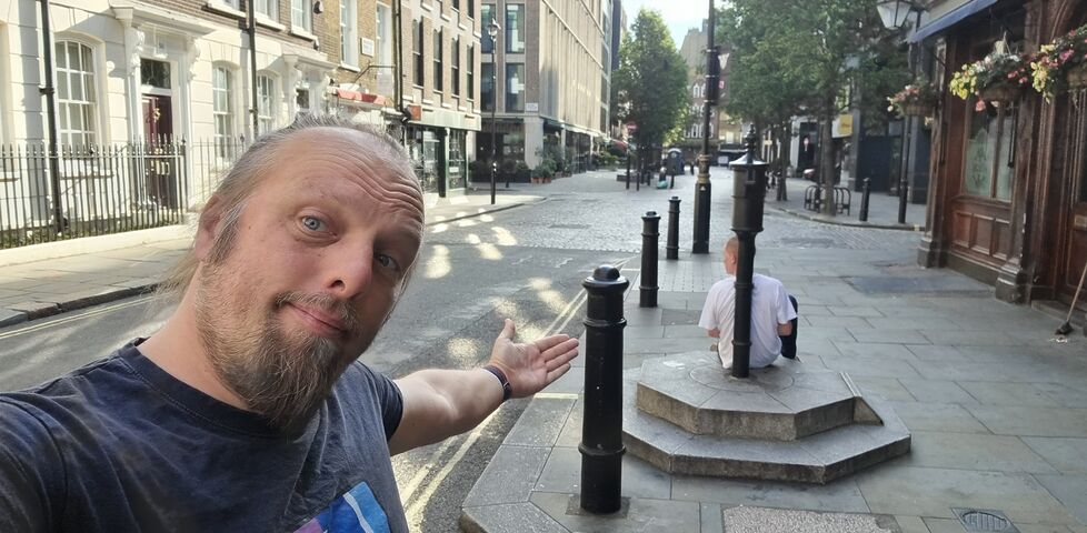 Dan stands on a London street, gesturing to an old water pump (against which a man reclines).