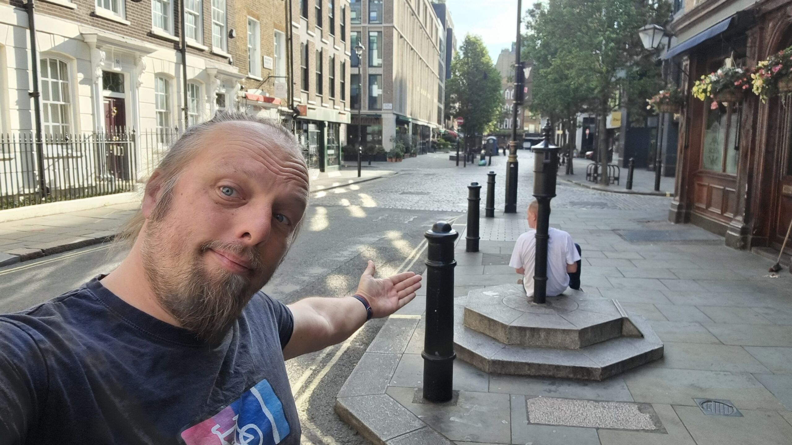 Full image of Dan stands on a London street, gesturing to an old water pump (against which a man reclines).
