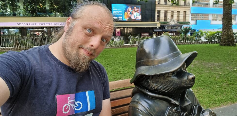 Dan sits on a park bench in Leicester Square, alongside a bronze sculpture of Paddington Bear.