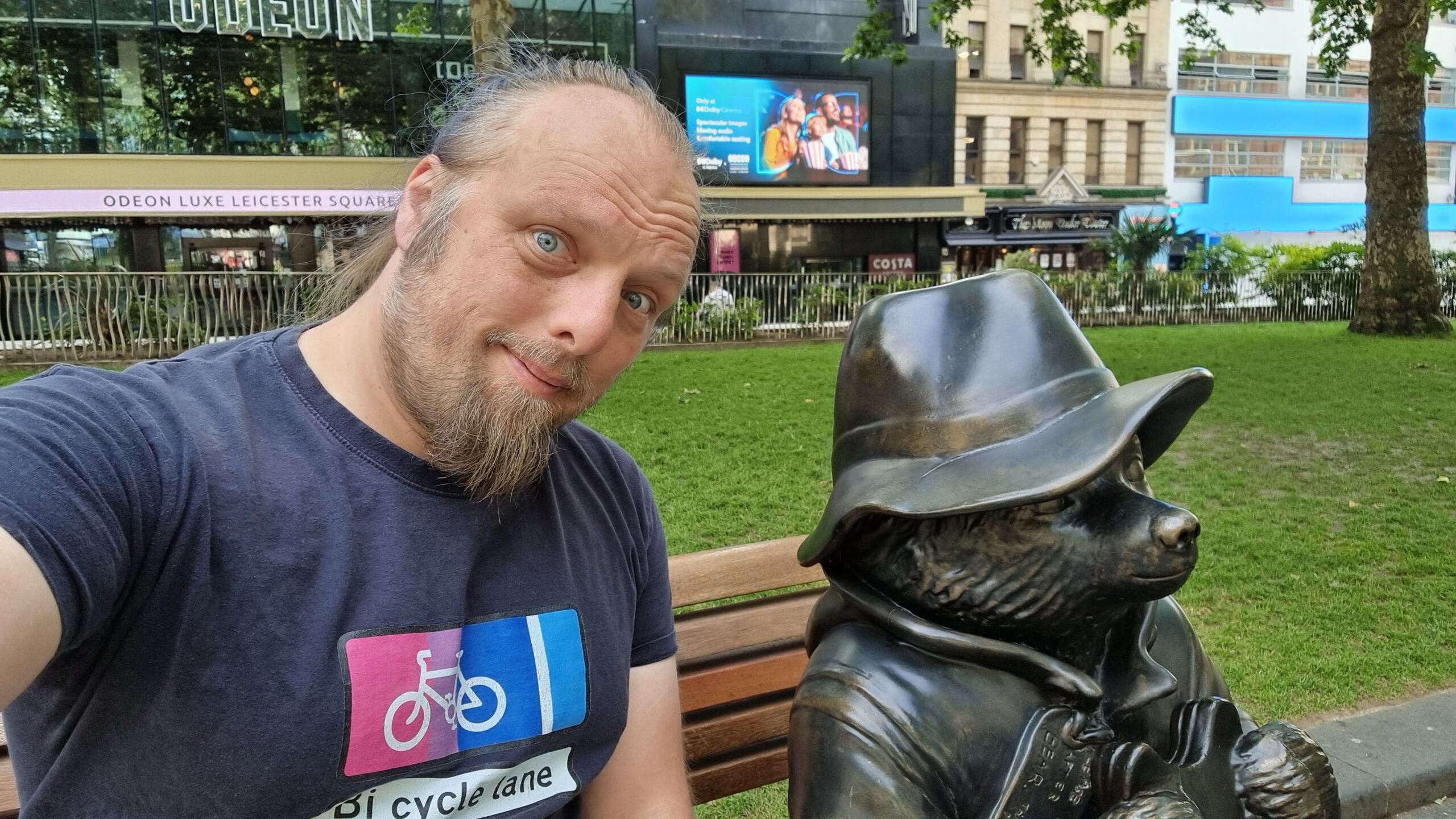 Full image of Dan sits on a park bench in Leicester Square, alongside a bronze sculpture of Paddington Bear.