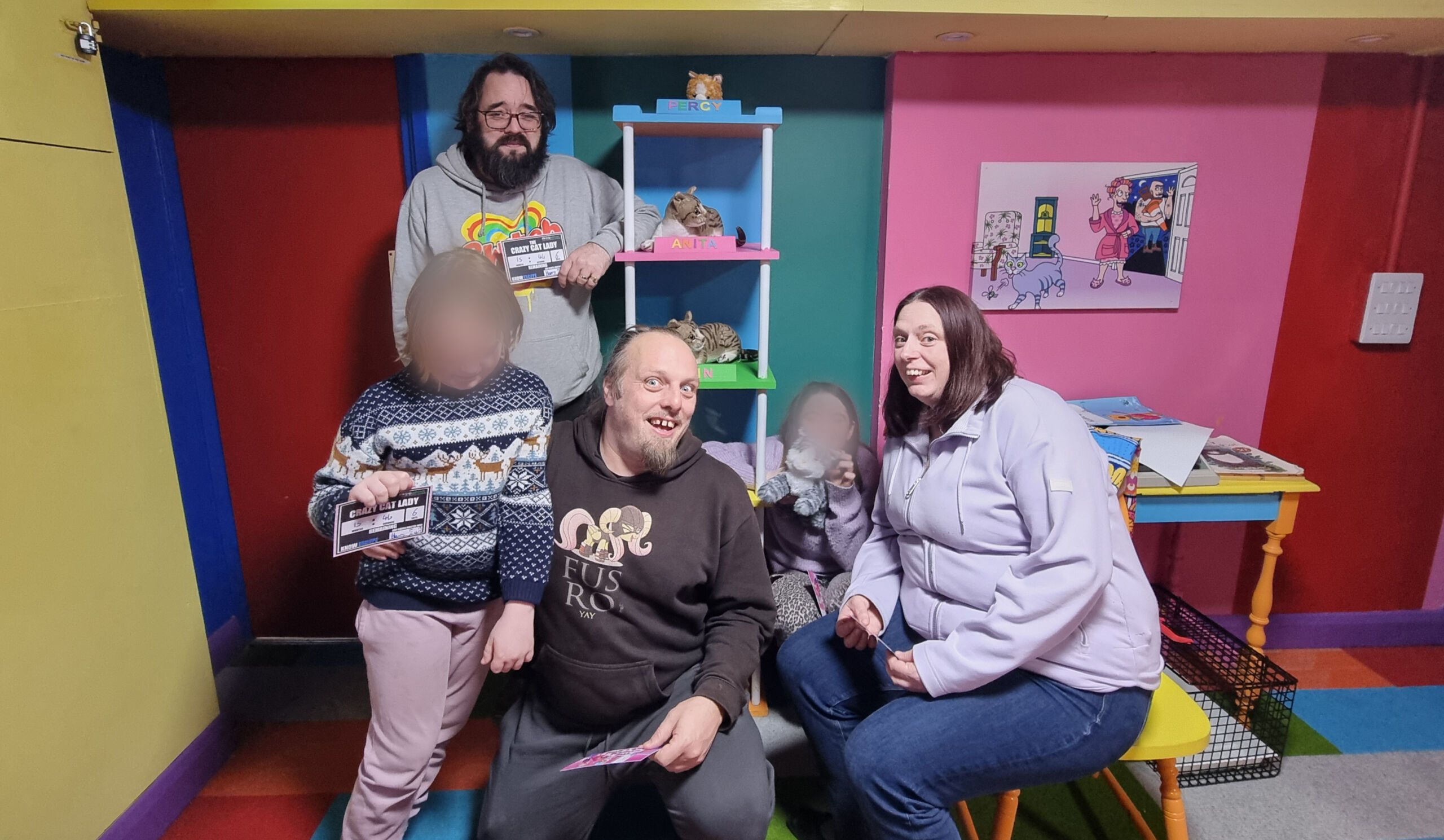 Full image of The same adults and children pose in a colourful escape room, with padlocked boxes and banks of light switches visible amongst cat toys.