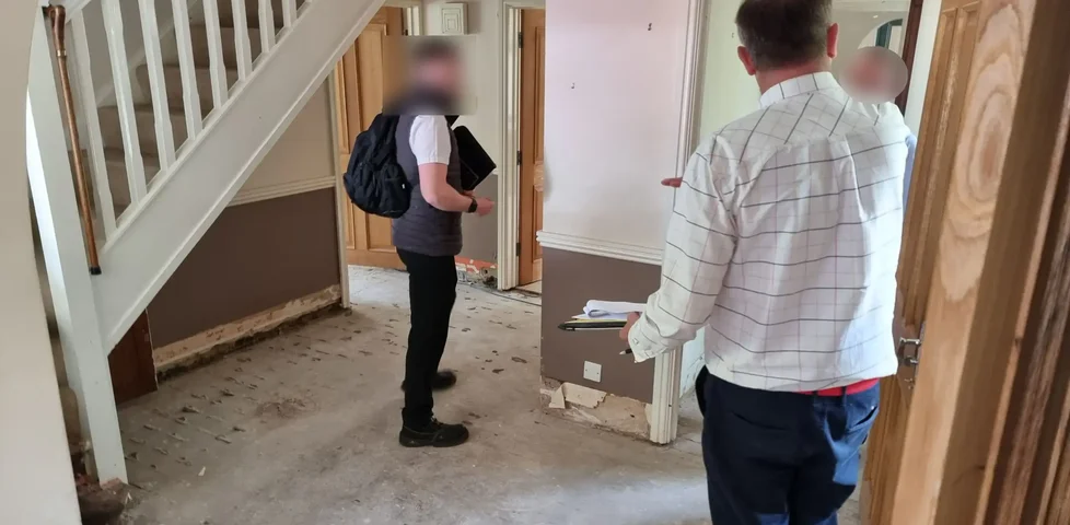 Several men stand on the bare concrete floor of a residential hallway with no furniture or skirting boards.
