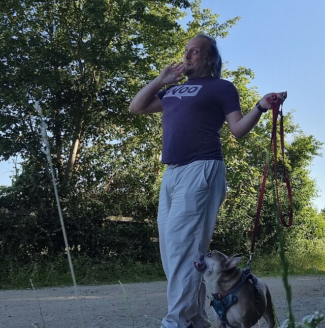 Dan poses with a coquetteish grin while walking a French Bulldog along a gravelled rural path.