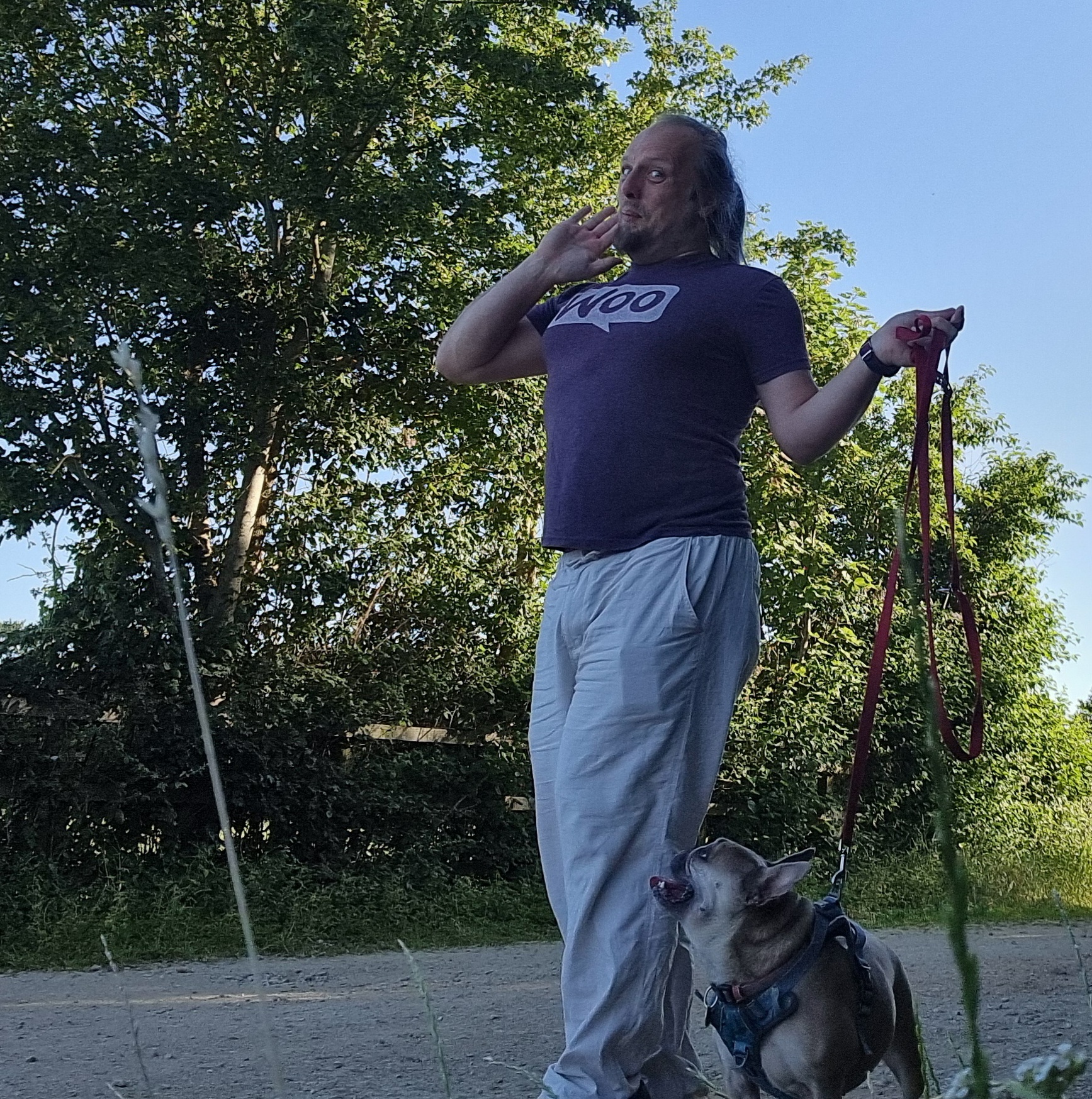 Full image of Dan poses with a coquetteish grin while walking a French Bulldog along a gravelled rural path.