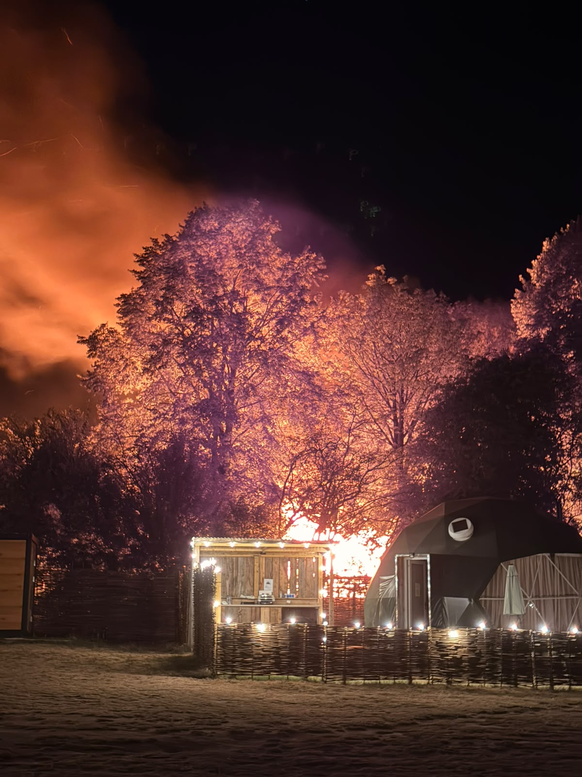 Full image of A static caravan is engulfed in flames behind a dome tent, at night.