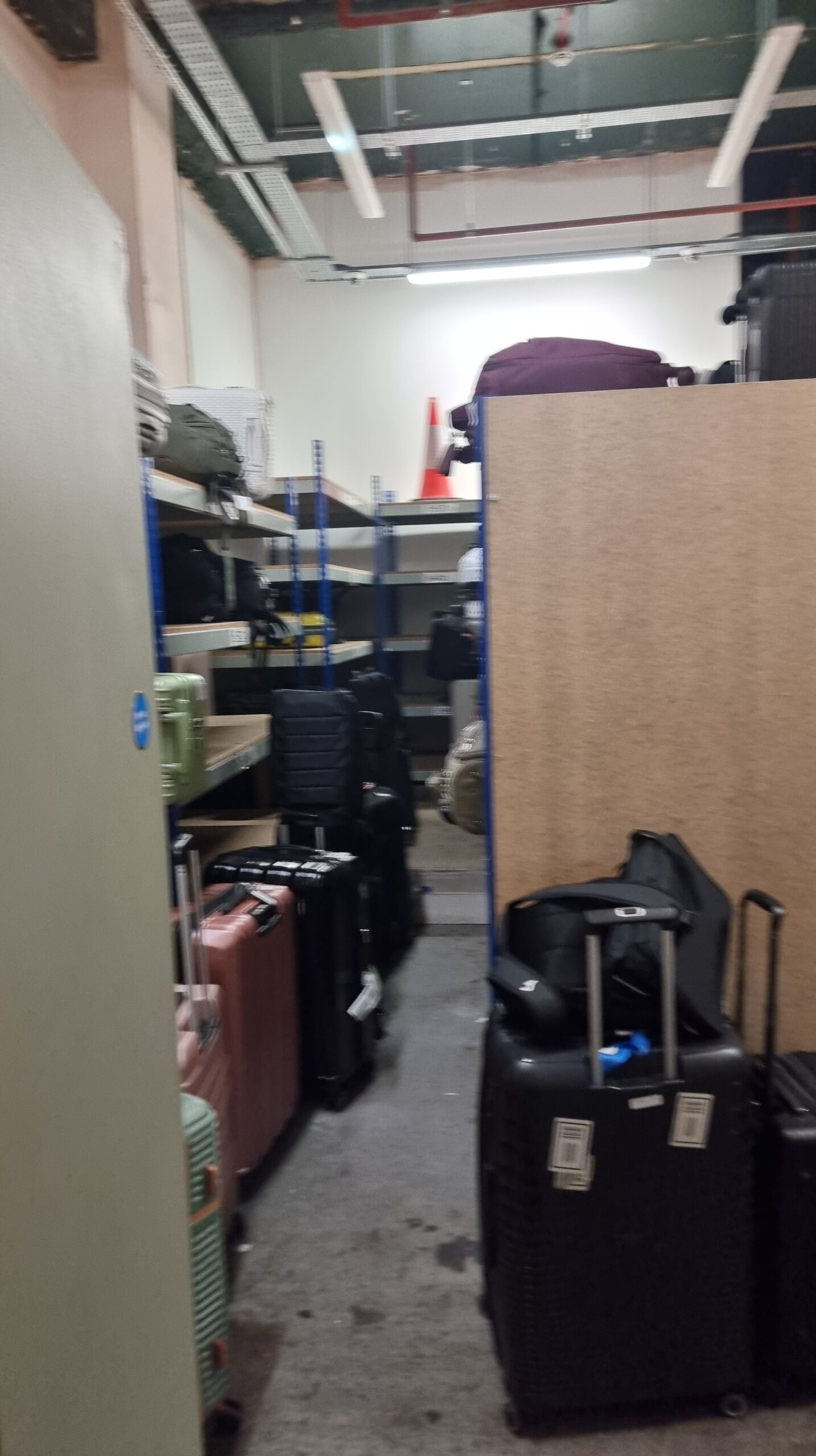 Full image of A cluttered left-luggage room with many suitcases stacked on shelves... and atop one set of shelves, a traffic cone.