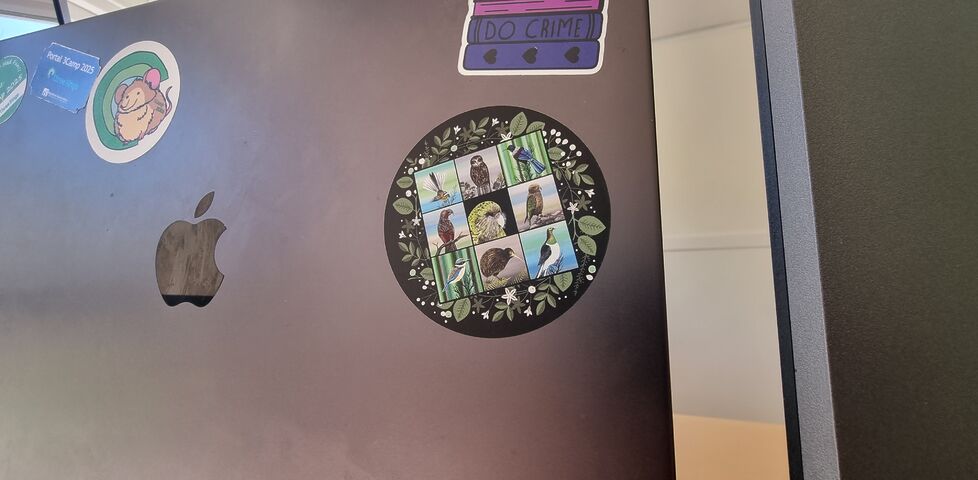 Circular sticker featuring nine New Zealand birds, stuck to a laptop under a sticker that says 'read gay; do crime'.