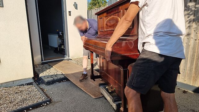 Two men move an upright piano across a driveway and through a door, with the help of a small trolley and some boards.