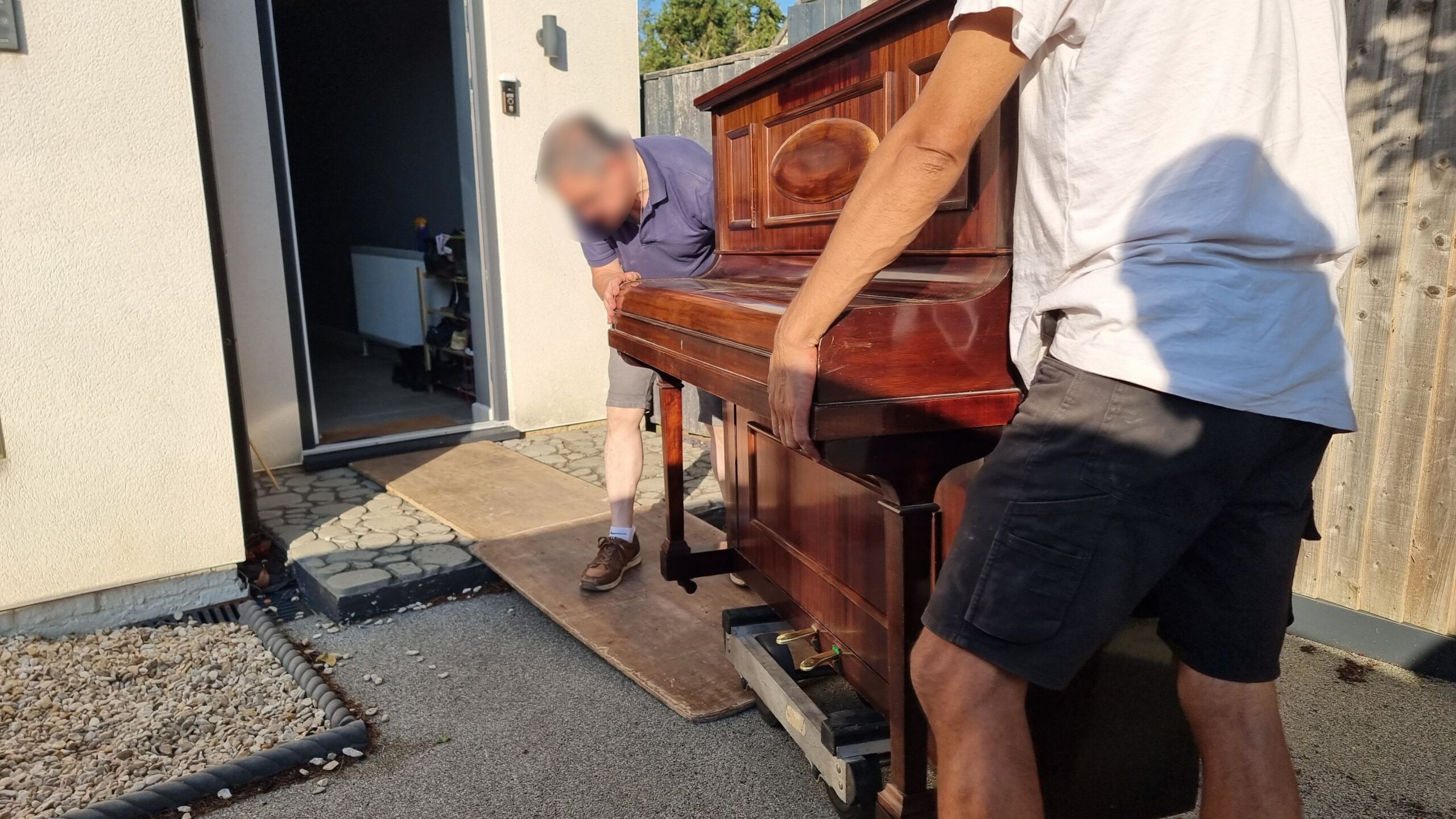 Full image of Two men move an upright piano across a driveway and through a door, with the help of a small trolley and some boards.