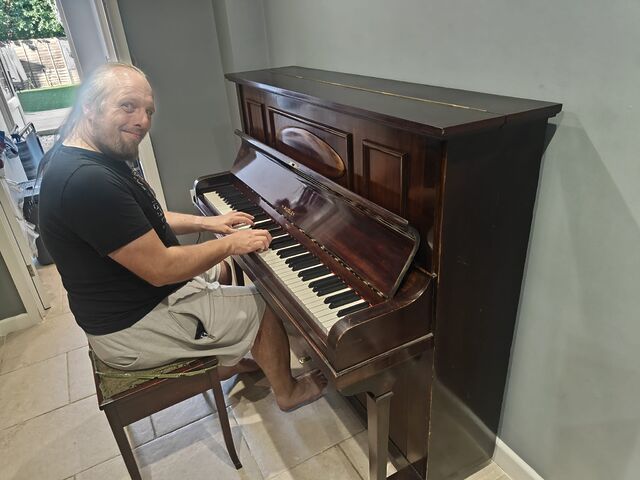 Dan, a white man with a beard and a blue-dyed ponytail, sits in a plain hallway at a polished upright piano, looking happy.