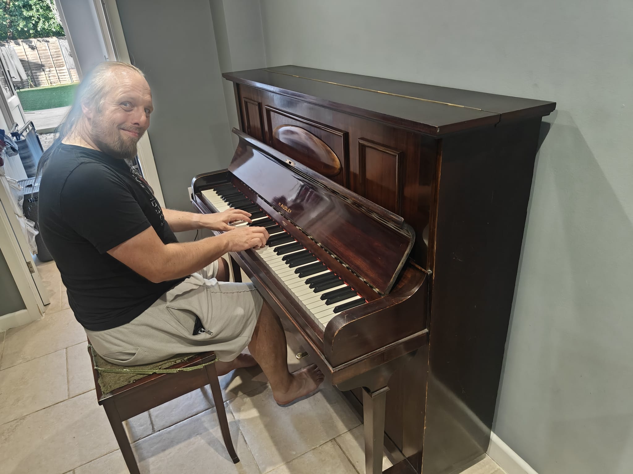 Full image of Dan, a white man with a beard and a blue-dyed ponytail, sits in a plain hallway at a polished upright piano, looking happy.