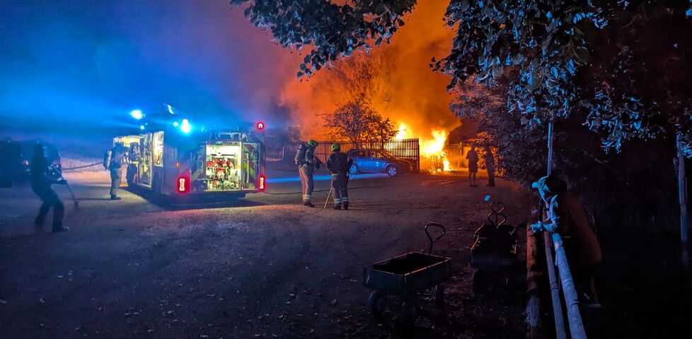 Firefighters pull hoses from a fire engine towards the inferno of a flaming cabin.